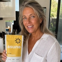 A blonde woman in her mid-40s, holding a yellow and white package of 'Asuno Cummies' dietary supplement, smiling in a well-lit kitchen with large windows in the background