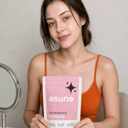 Asian woman in an orange tank top holding a pink bag labeled Asune Skin Brightening Gummies, smiling softly against a neutral background