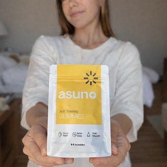 A woman in a white top holding a packet labeled Su No, a self tanning gummy product for one week use. The packet is white with a yellow strip, featuring the Su No logo and a product icon indicating suitability for sensitive skin.